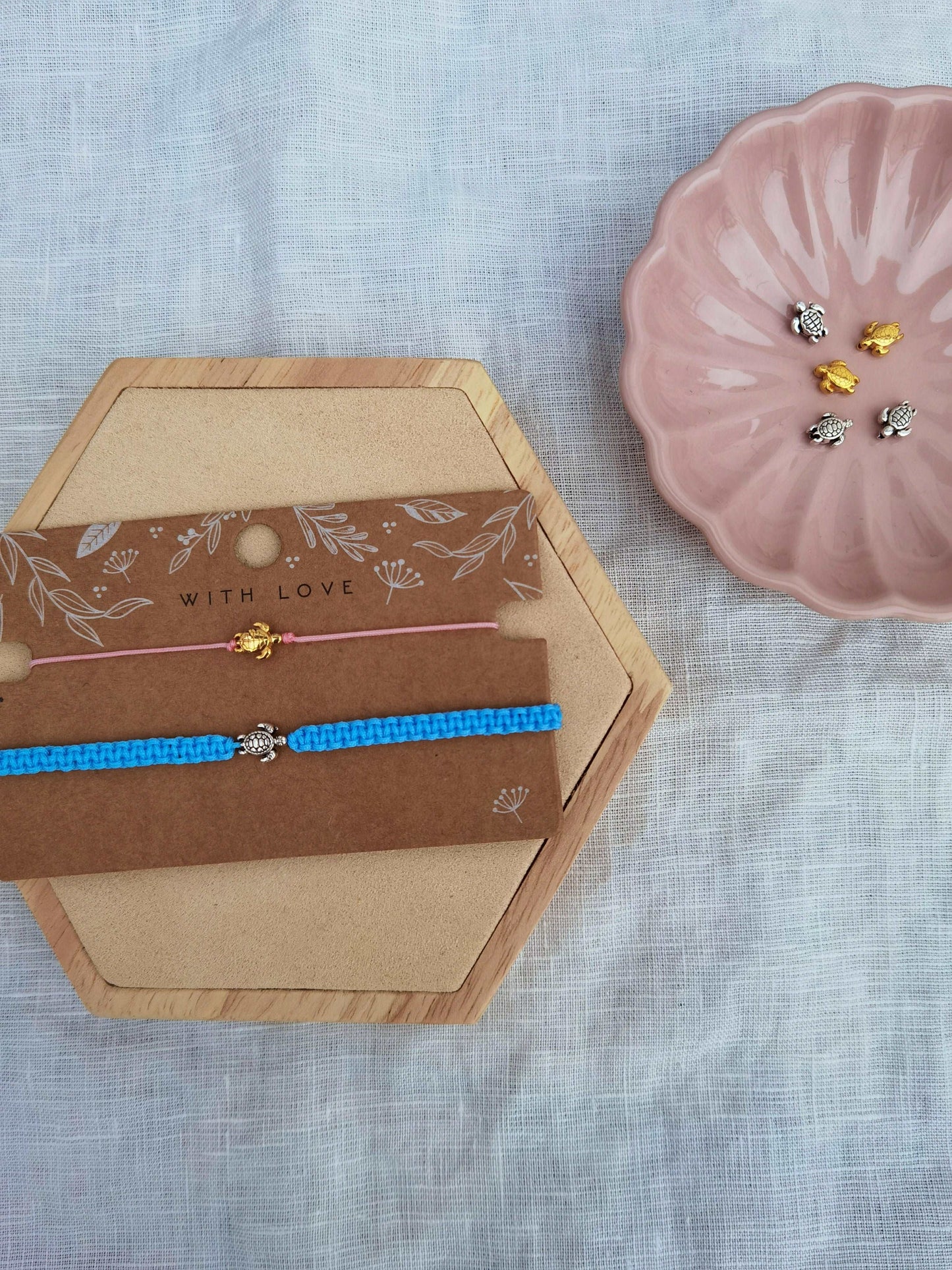 Zwei Schildkröten Armbänder auf einer braunen Schmuckkarte. Oberes Armband rosa mit goldener Schildkröte. Unteres Armband hellblau mit silberner Schildkröte. Daneben eine rosa Schale mit weiteren goldenen und silbernen Schildkröten Perlen