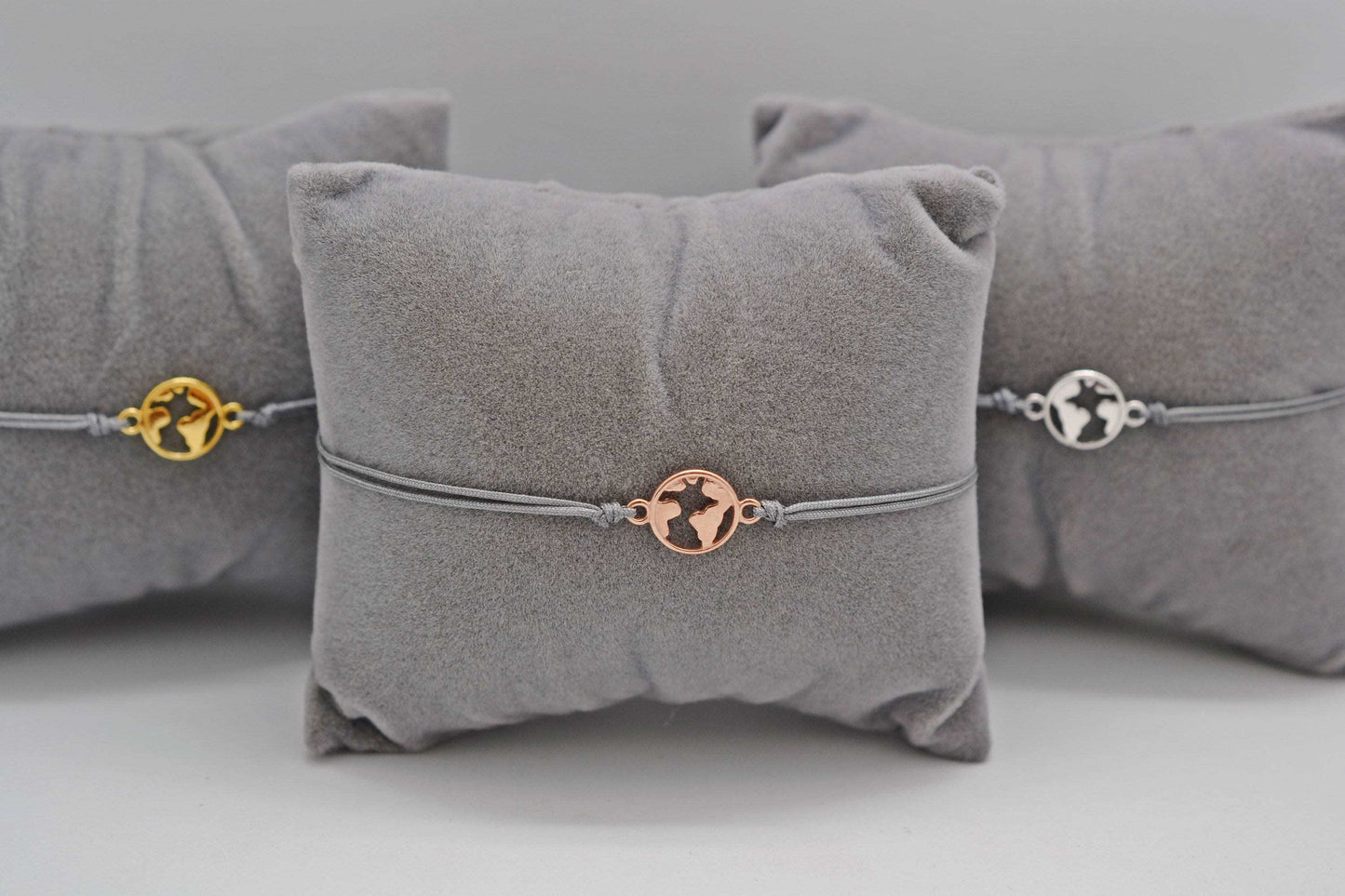 Drei Weltkugel Armbänder in roségold, gold und silber mit grauem Band auf grauen Schmuckkissen