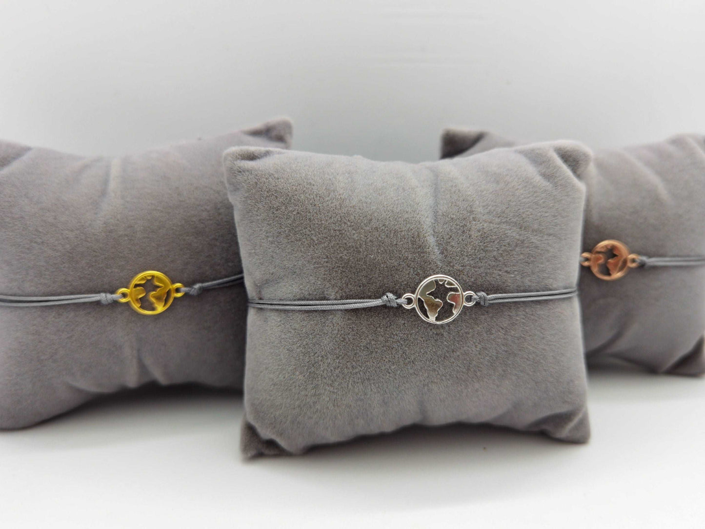 Mini Weltkarte Armband in silber, gold und roségold mit silbernem Band jeweils auf einem grauen Schmuckkissen