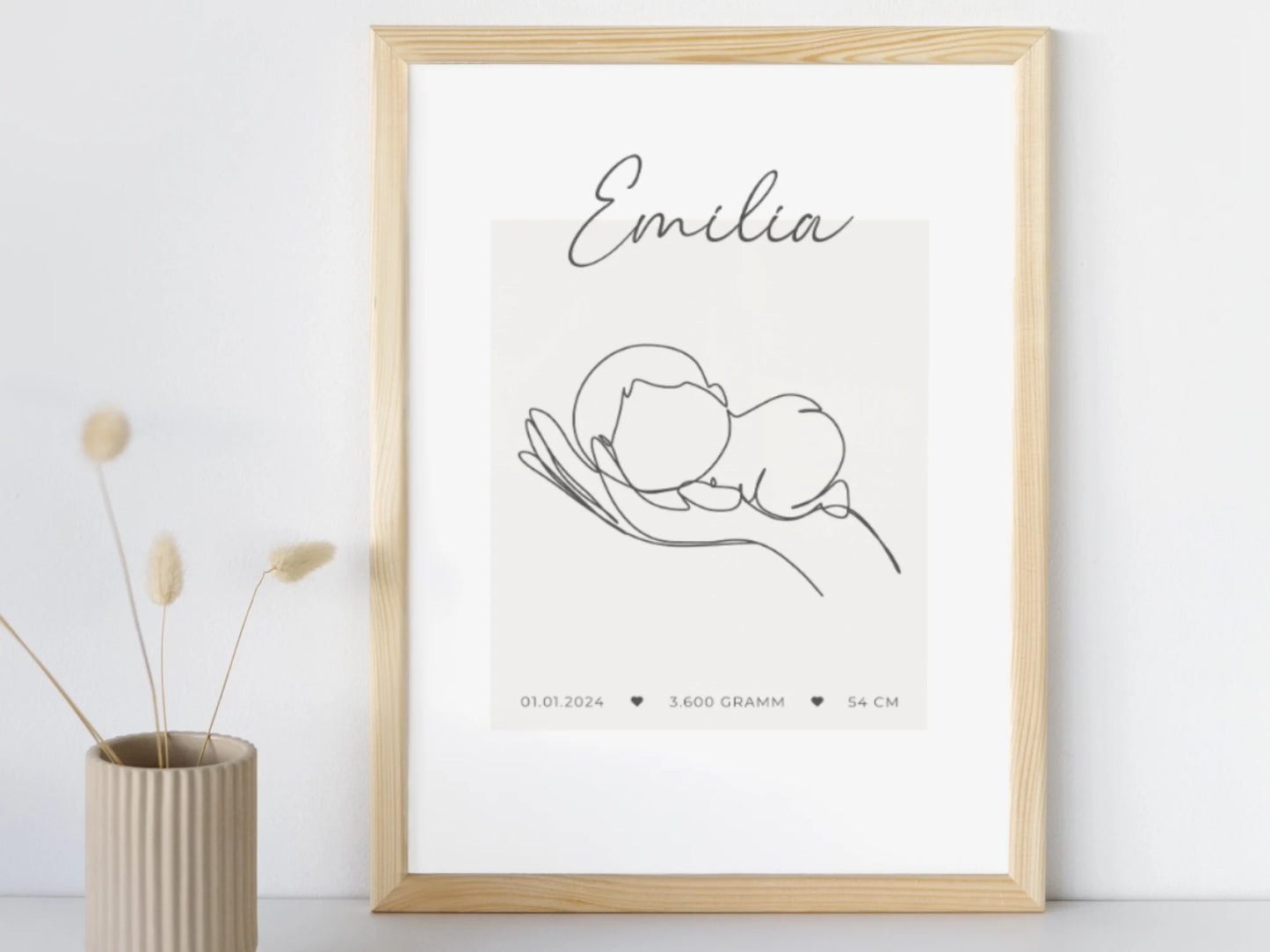 Geburtsposter mit einer Hand, die ein Baby hält. Darüber der Name "Emilia", darunter das Geburtsdatum, das Geburtsgewicht und die Geburtsgröße. Poster in einem Holzrahmen an einer Wand lehnend