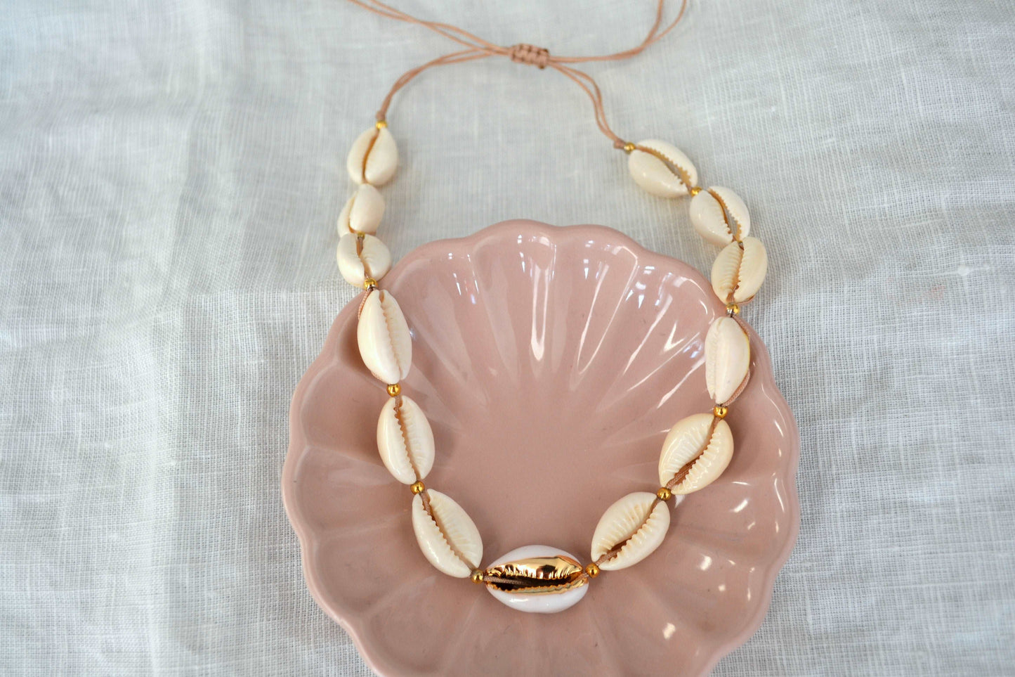 Muschel Kette mit goldenen Akzenten und beigem Band in einer rosa Schmuckschale und vor weißem Hintergrund