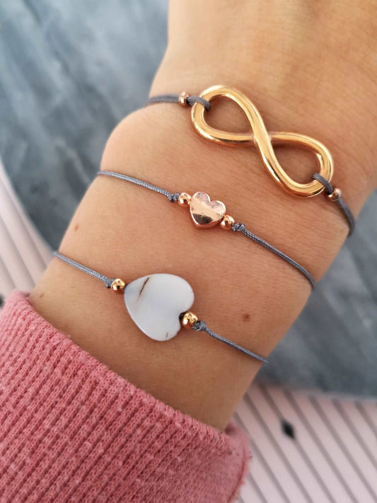 Tragefoto drei verschiedene Armbänder mit roségoldenen Elementen und grauem Band. Unteres Armband Muschel Herz Armband, in der Mitte ein Herz Armband und oben ein Unendlichkeitszeichen Armband
