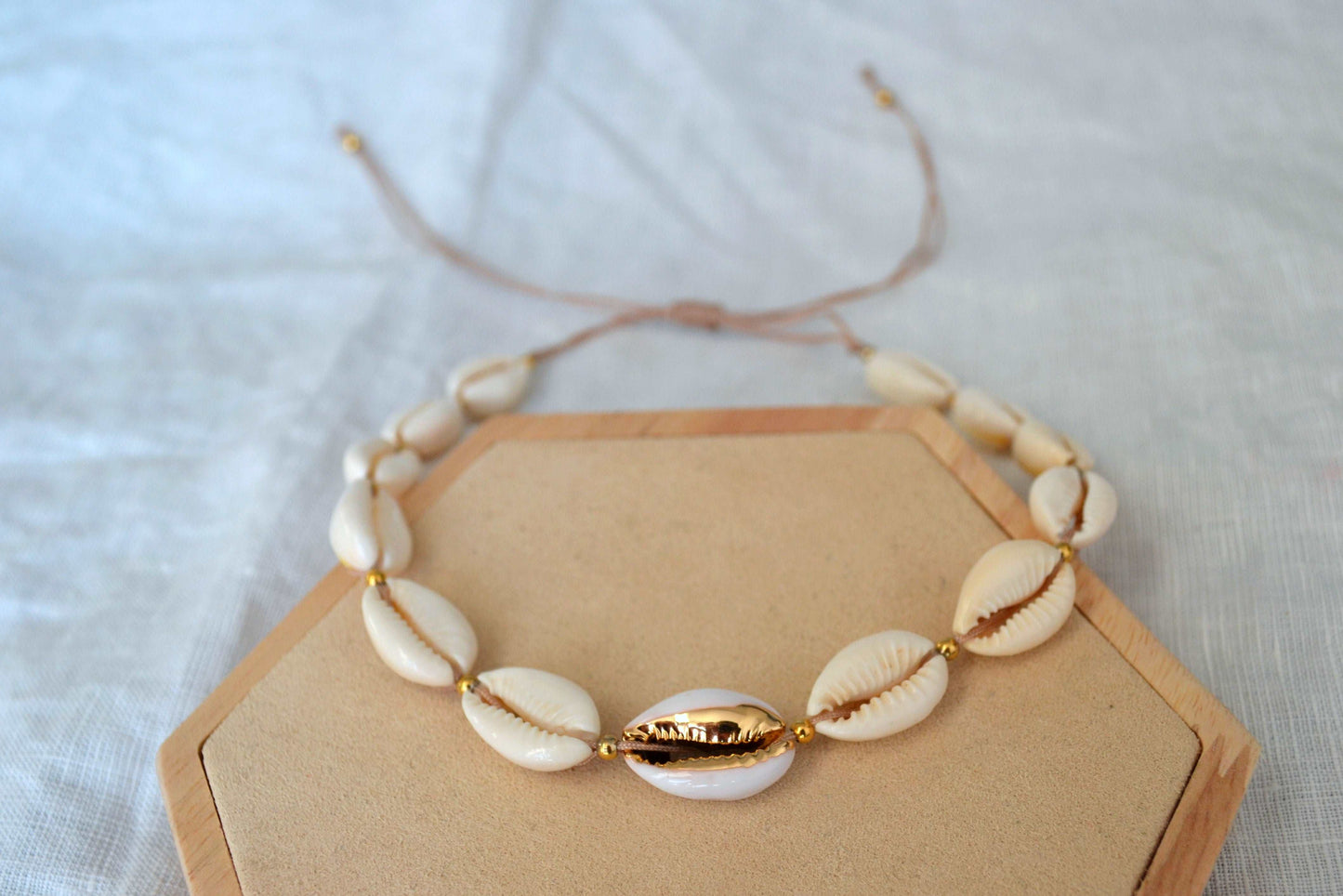 Muschel Choker mit goldenen Akzenten und einem beigen Band auf einem Holz Sechseck vor weißem Hintergrund