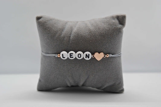 Personalisiertes Buchstaben Armband mit dem Namen Leon, einem roségoldenen Herz und grauem Band. Fotografiert auf einem grauen Schmuckkissen