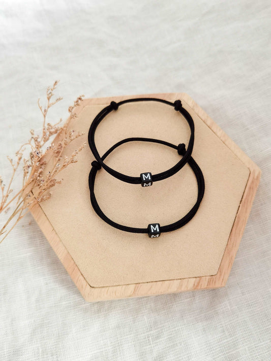 Zwei schwarze personalisierte Partnerarmbänder aus Segeltau beide mit Buchstaben "M" auf einem Holz Sechseck. Foto im Hochformat