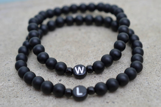 Zwei personalisierte Partnerarmbänder mit matt schwarzen Perlen liegen übereinander auf einem Steinboden