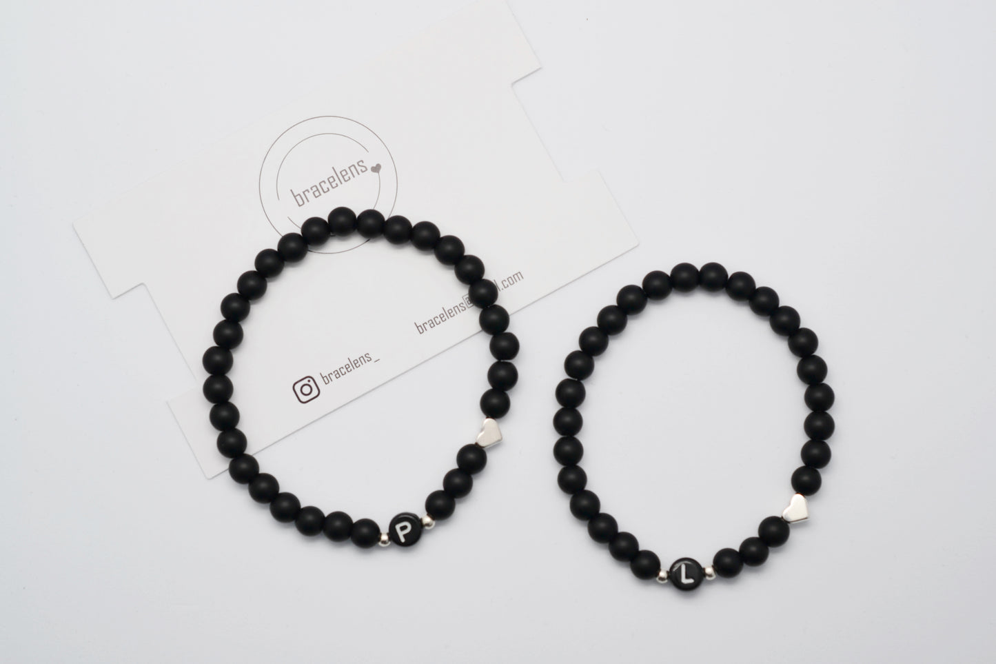 Zwei schwarze personalisierte Partnerarmbänder mit Initiale liegen nebeneinander vor weißem Hintergrund