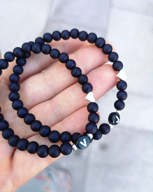 Zwei personalisierte schwarze Partnerarmbänder mit einem Buchstaben nach Wahl und silbernen Perlen und Herzen liegen auf einer Hand