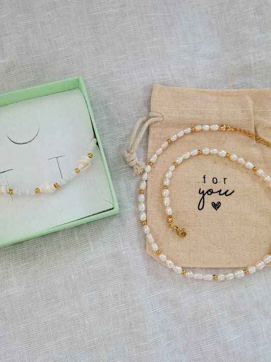 Eine weiß goldene Muschel Fußkette in einer mintgrünen Schmuckschachtel liegt neben einer weißen Süßwasserperlenkette mit goldenen Akzenten, die zu einer Schnecke aufgerollt ist