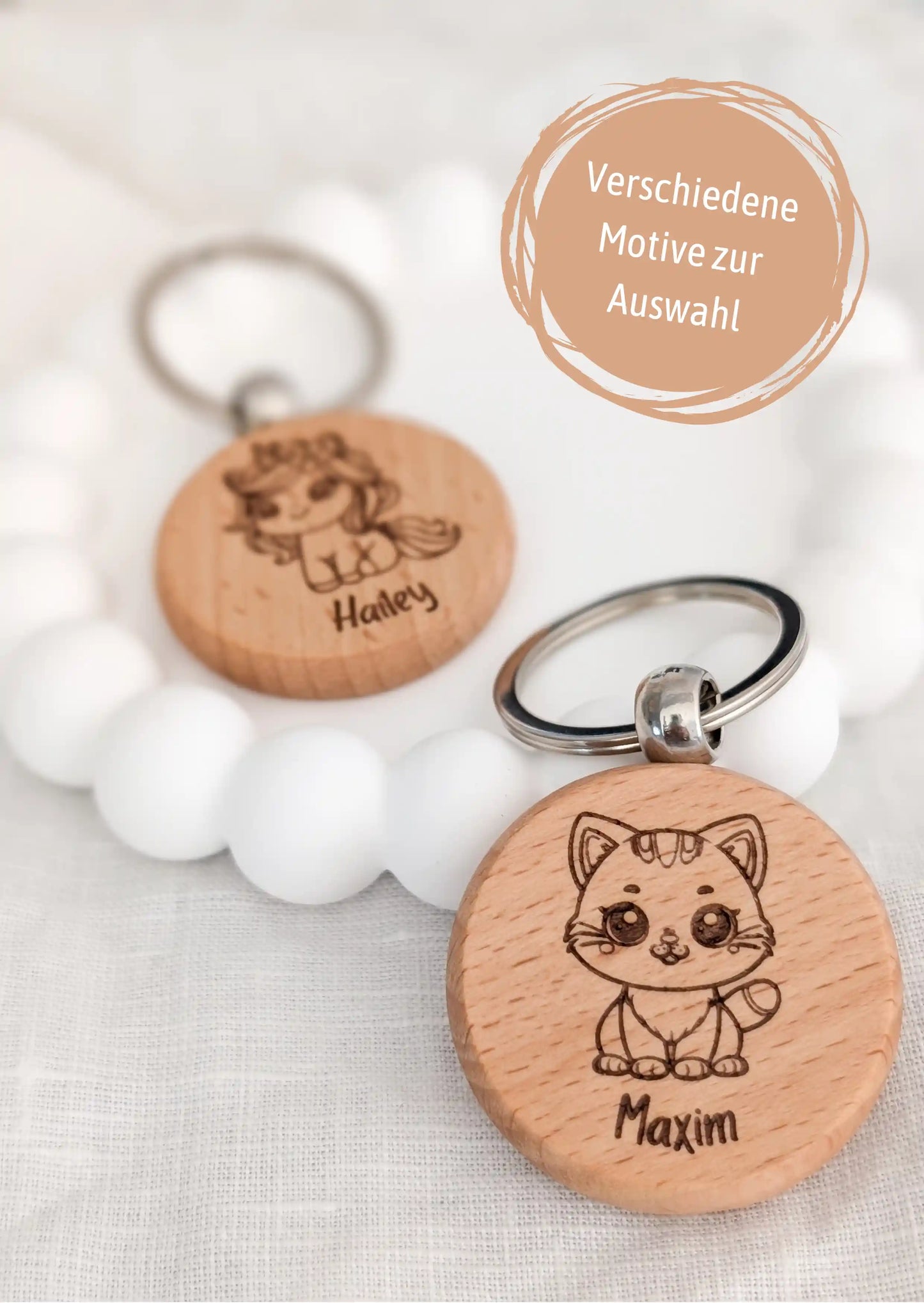 Im Vordergrund ein personalisierter Schlüsselanhänger für Kinder mit einer süßen Katze und dem Namen Maxim. Dahinter auf einem weißen Untersetzer ein Schlüsselanhänger mit einem Einhorn und dem Namen Hailey