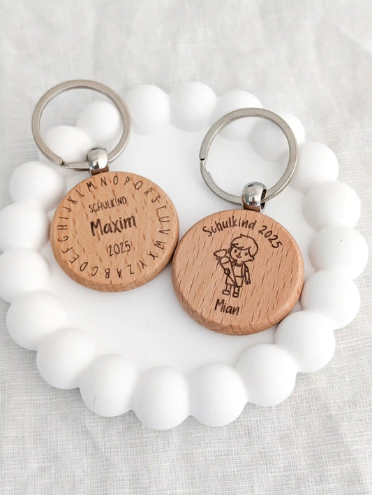 Zwei personalisierte Schlüsselanhänger Schulkind als Geschenk zur Einschulung liegen nebeneinander auf einem weißen Untersetzer. Links "Schuldkind Maxim 2025" und drum herum das Alphabet eingraviert. Rechts "Schulkind 2025" darunter ein Junge mit Schultüte und darunter der Name "Mian"