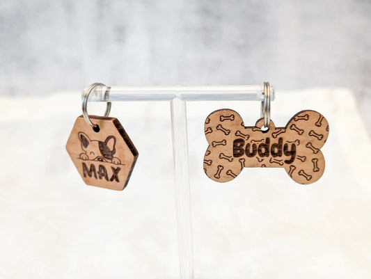 Zwei personalisierte Hundemarken aus Holz hängen an einem durchsichtigen Schmuckständer. Links ein sechseckiger Anhänger mit einem Hund und "Max" und rechts ein knochenförmiger Anhänger mit kleinen Knochen im Hintergrund und dem Namen "Buddy" im Vordergrund
