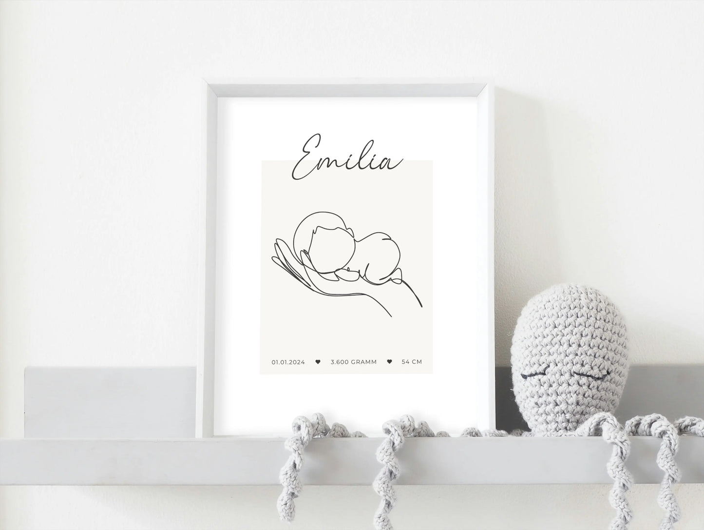 Geburtsposter mit einer Hand, die ein Baby hält. Darüber der Name "Emilia", darunter das Geburtsdatum, das Geburtsgewicht und die Geburtsgröße. Poster in einem weißen Rahmen auf einem Regal. Daneben ein gehäkelter Oktopus in grau