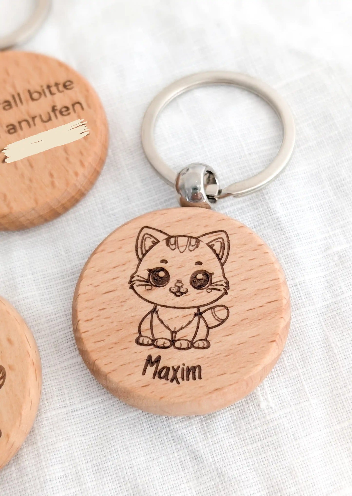 Ein personalisierter Schlüsselanhänger für Kinder aus Holz mit einer süßen Katze und dem Namen Maxim. Links daneben im Anschnitt zwei weitere Schlüsselanhänger