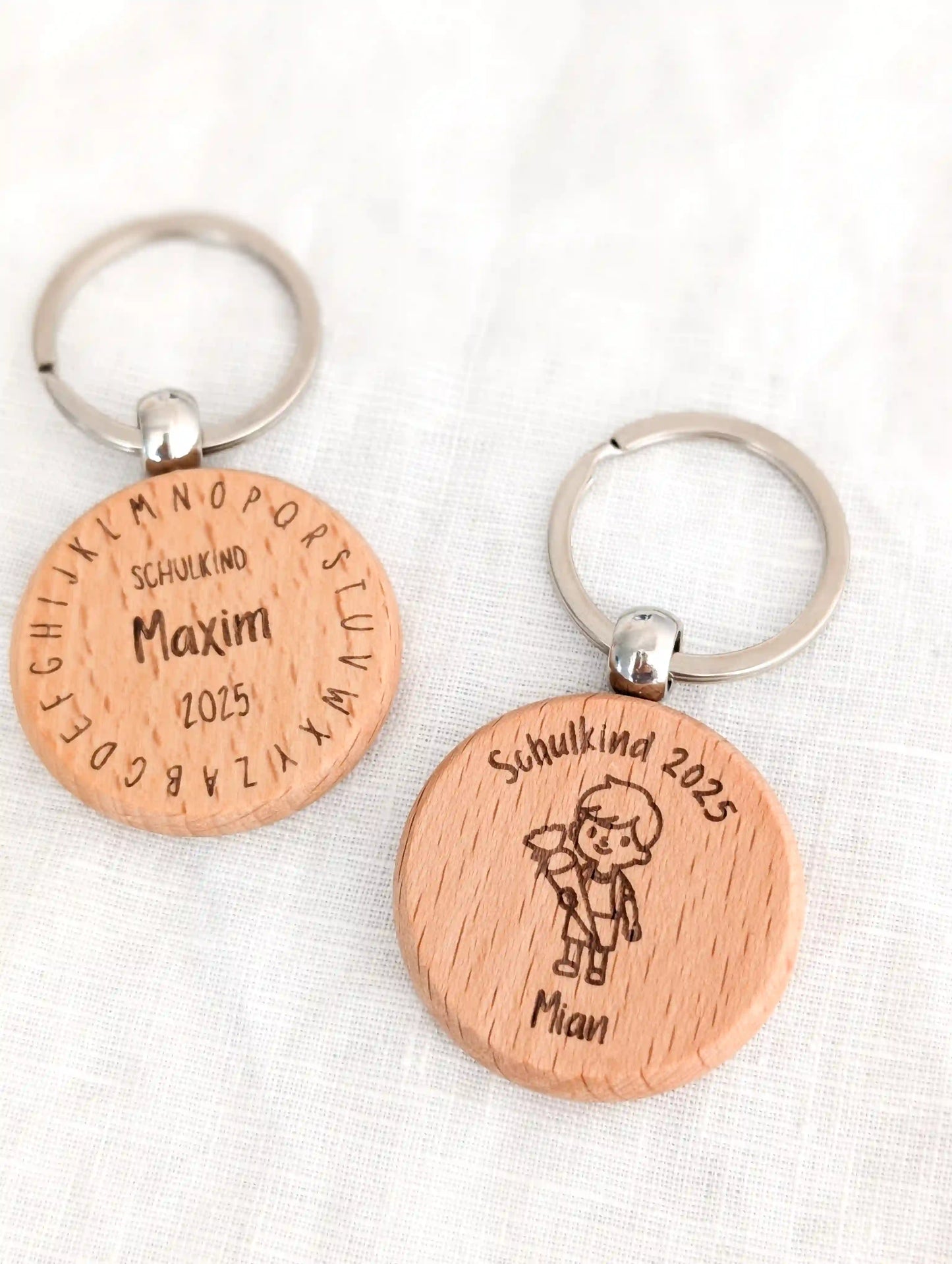 Zwei personalisierte Schlüsselanhänger Schulkind aus Holz liegen nebeneinander vor weißen Hintergrund. Links "Schuldkind Maxim 2025" und drum herum das Alphabet eingraviert. Rechts "Schulkind 2025" darunter ein Junge mit Schultüte und darunter der Name "Mian"
