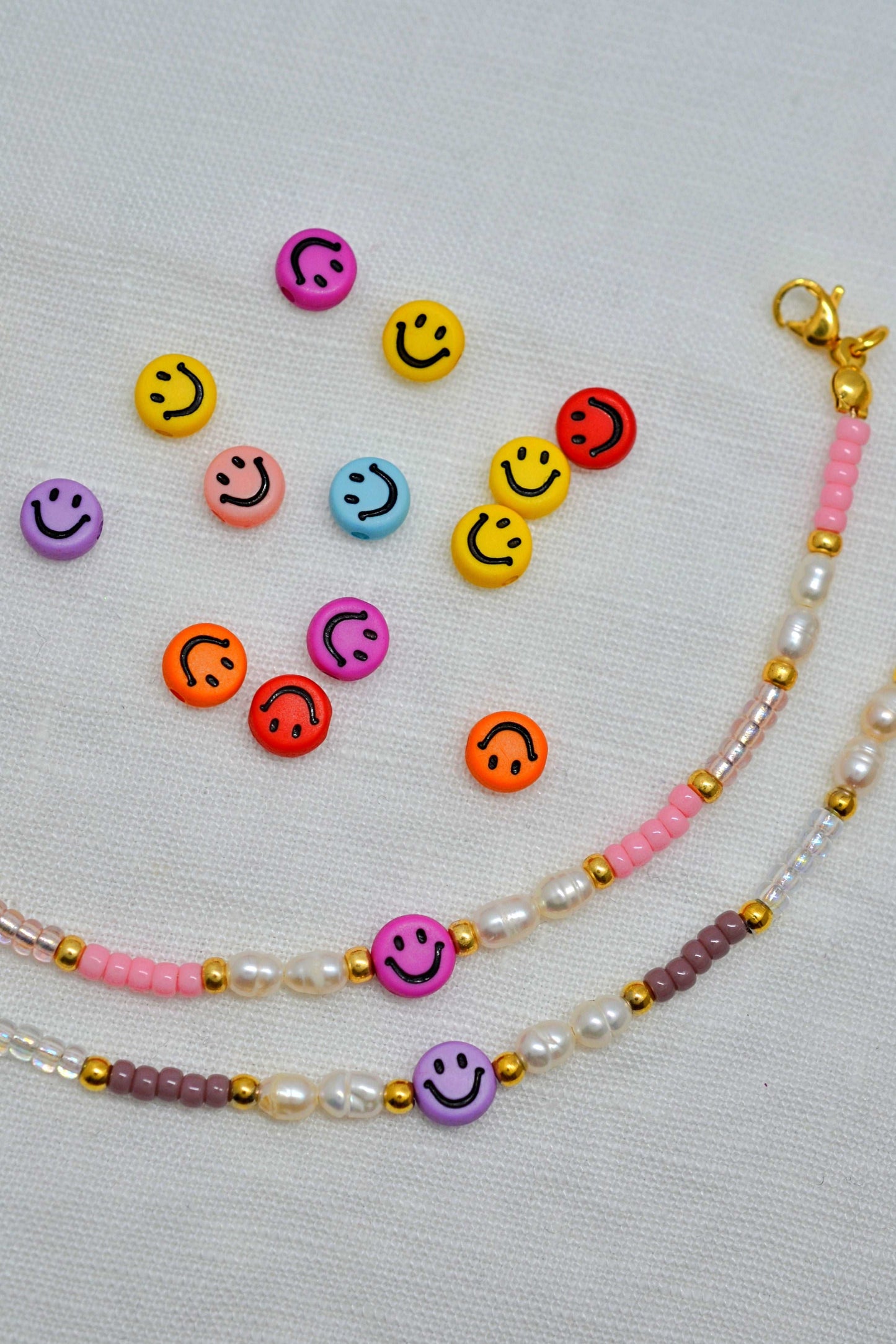 Zwei Gute Laune Armbänder mit Smileys und Süßwasserperlen in lila und rosa liegen voreinander auf weißem Hintergrund. Dahinter weitere bunte Smiley Perlen