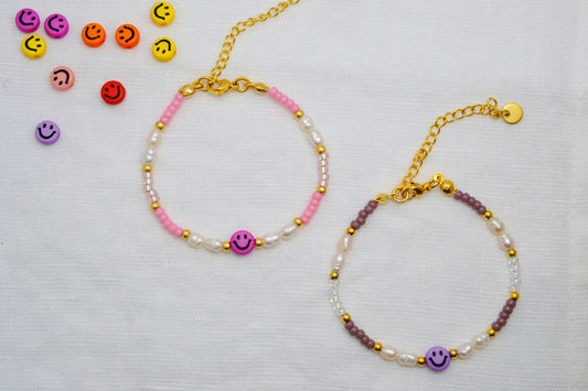 Ein rosa Smiley Armband mit Süßwasserperlen und ein lila Smiley Armband mit Süßwasserperlen liegen nebeneinander auf weißem Hintergrund. Daneben weitere bunte Smiley Perlen