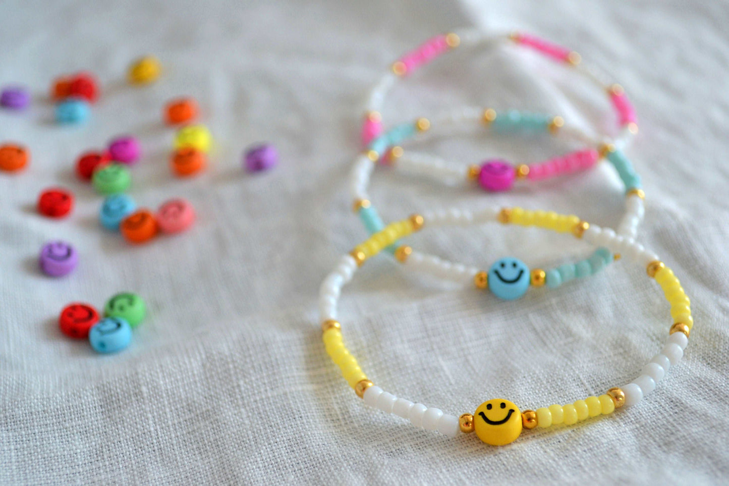 Buntes Perlenarmband mit Smiley in gelb, blau und rosa übereinander gelegt. Daneben weitere bunte Smileyperlen