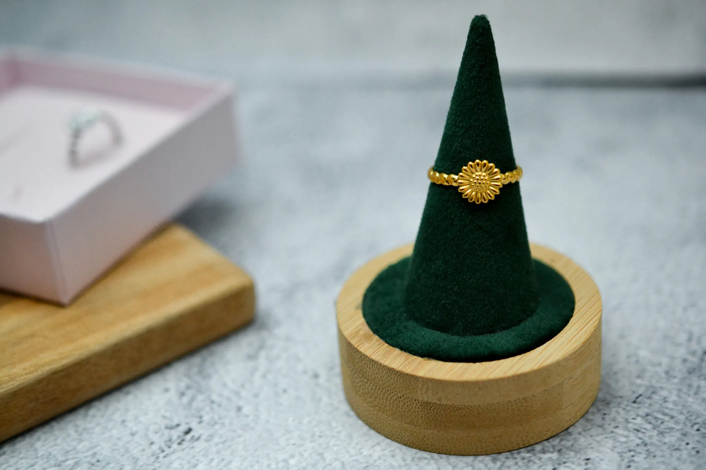 Goldener Sonnenblume Ring auf einem samtgrünen Ringhalter. Im Hintergrund eine rosa Schmuckschachtel mit demselben Ring in silber
