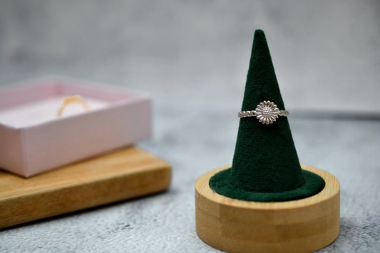 Silberner Sonnenblume Ring auf einem samtgrünen Ringhalter. Im Hintergrund unscharf eine rosa Schmuckschachtel mit demselben Ring in gold dargestellt
