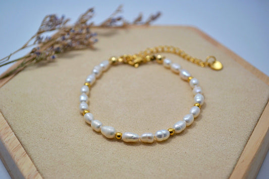 Ein weißes Süßwasserperlen Armband mit goldenen Perlen auf einem Holz Sechseck. Im Hintergrund ein Zweig getrocknete Blumen.