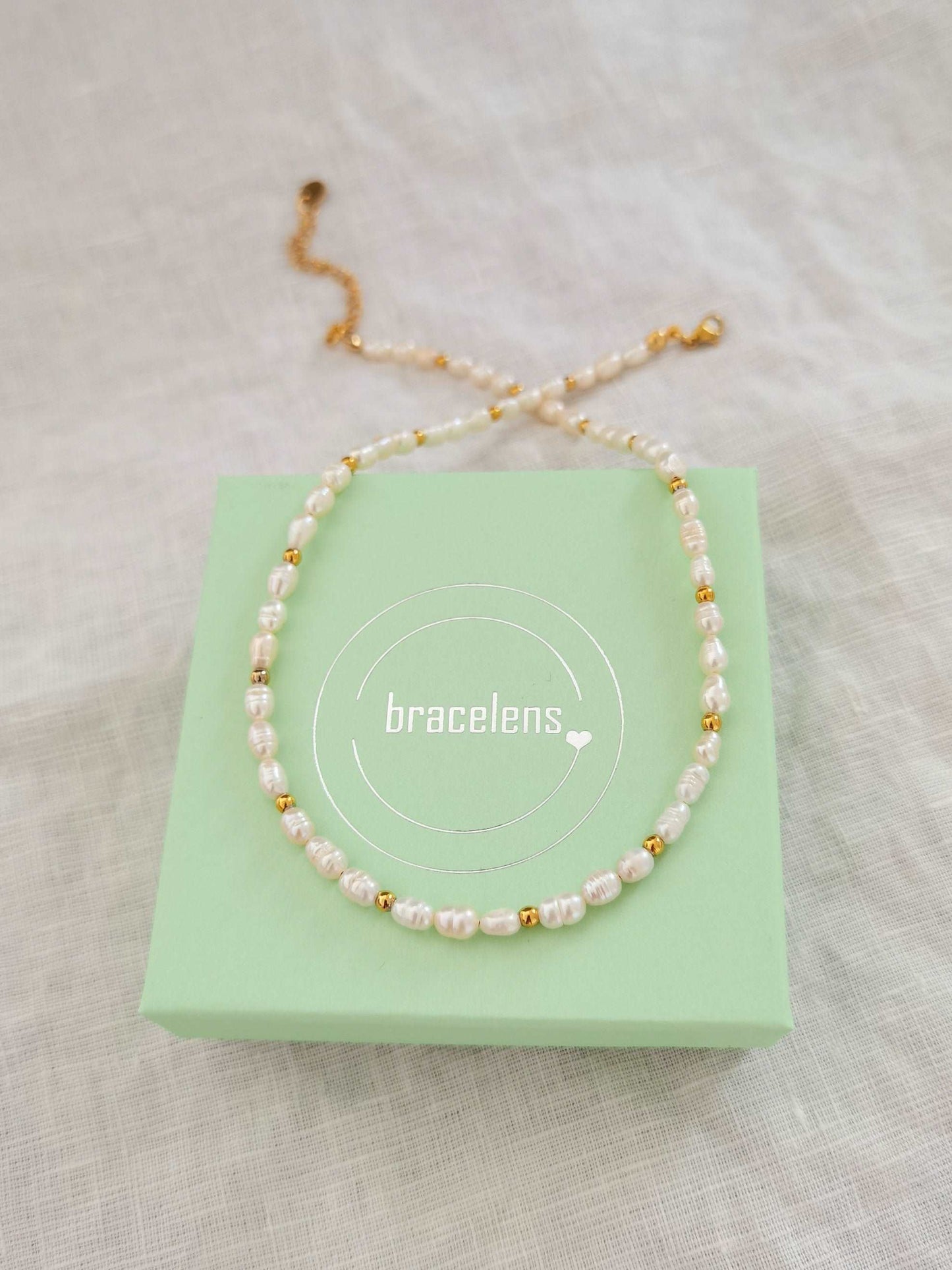 Eine Kette aus weißen Süßwasserperlen und kleinen goldenen Edelstahlperlen liegt auf einer mintgrünen Schmuckschachtel mit bracelens Logodruck