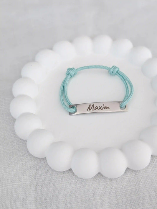 Ein mintgrünes Taufarmband für Kinder mit Gravur "Maxim" liegt auf einem weißen Untersetzer