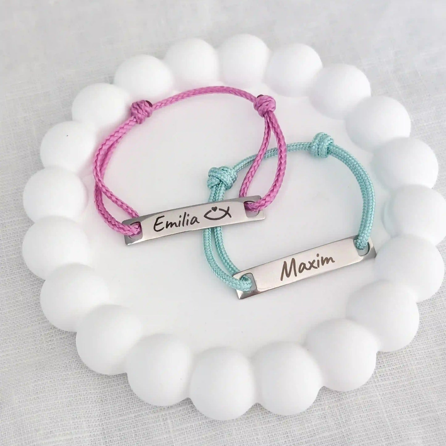 Zwei personalisierte Taufarmbänder für Kinder liegen auf einem weißen Untersetzer. Links pinkes Segeltau Armband mit Gravur "Emilia" und einem religiösen Fischzeichen. Rechts mintgrünes Taufarmband mit Gravur "Maxim"