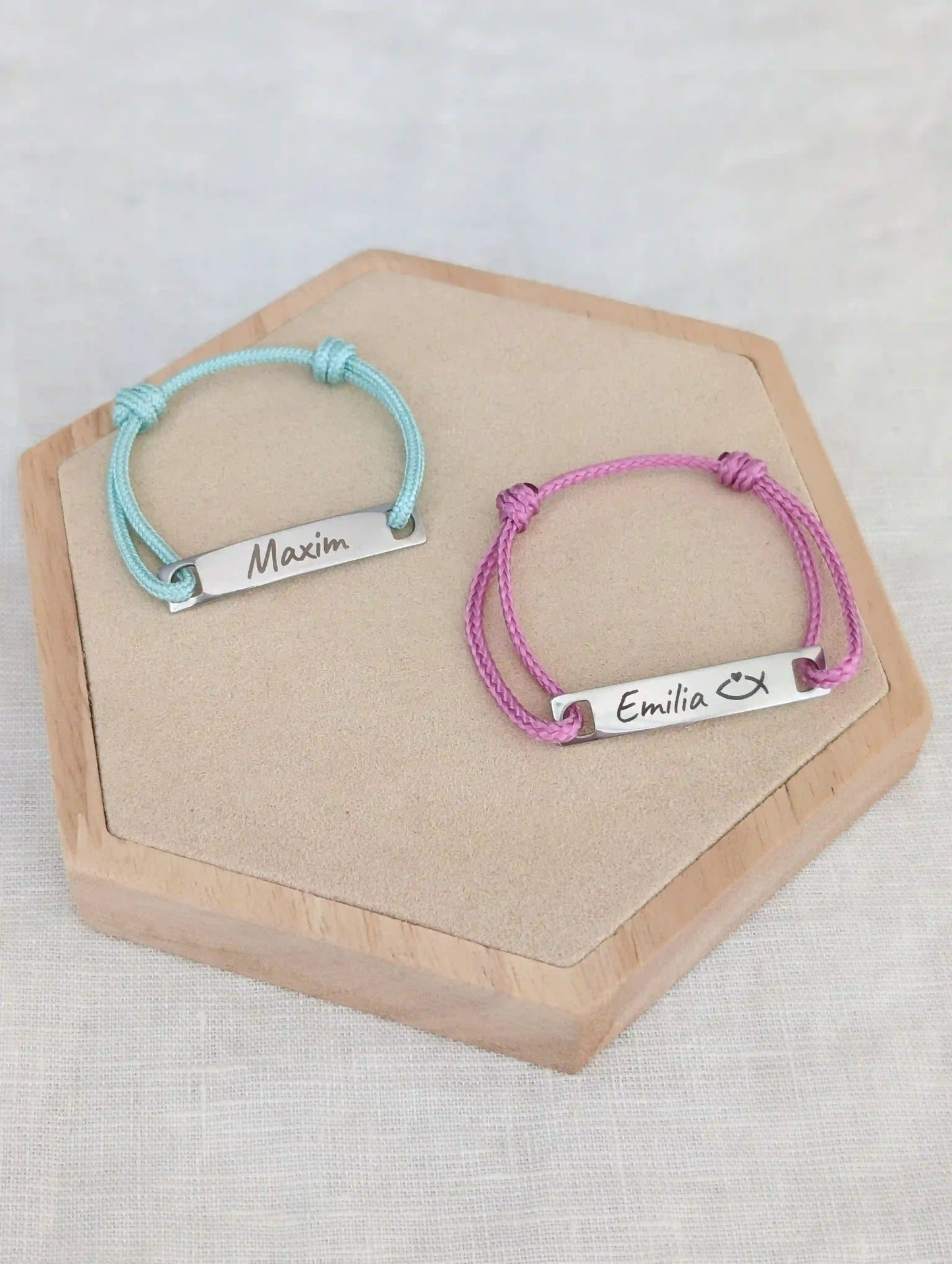 Zwei Taufarmbänder mit Namen liegen auf einem Holz Sechseck. Links mintgrünes Taufarmband mit Gravur "Maxim" rechts rosafarbenes Armband mit "Emilia" und einem religiösen Fisch Symbol