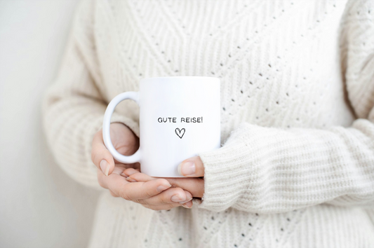 Weiße Tasse mit Aufschrift "Gute Reise!" und einem Herz wird von einer Frau in einem hellen Wollpulli in der Hand gehalten