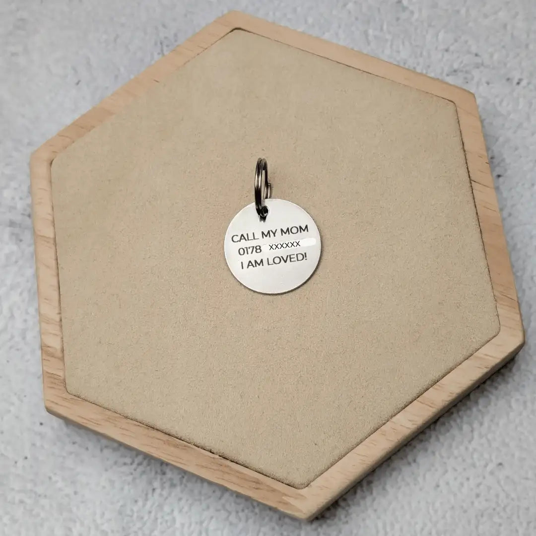 Silberne Hundemarke liegt mit der Rückseite nach oben auf einem Holz Sechseck. Gravur "Call My Mom, Handynummer, I am loved!"