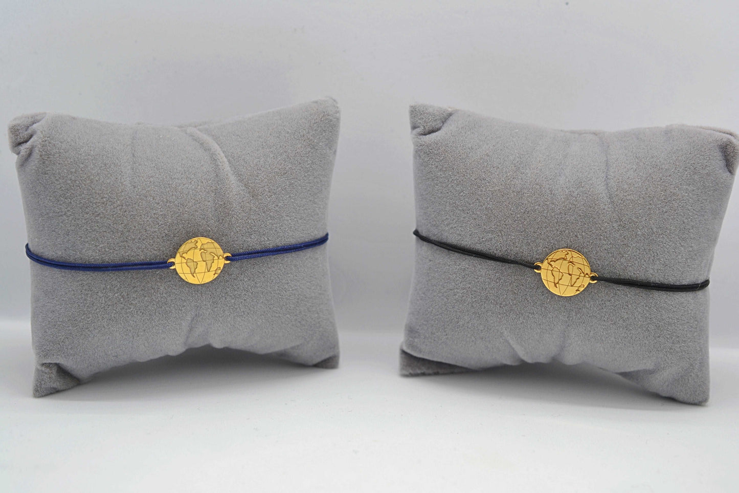 Zwei goldene Weltkarte Armbänder eins mit blauem und eins mit schwarzem Band jeweils auf einem grauen Schmuckkissen