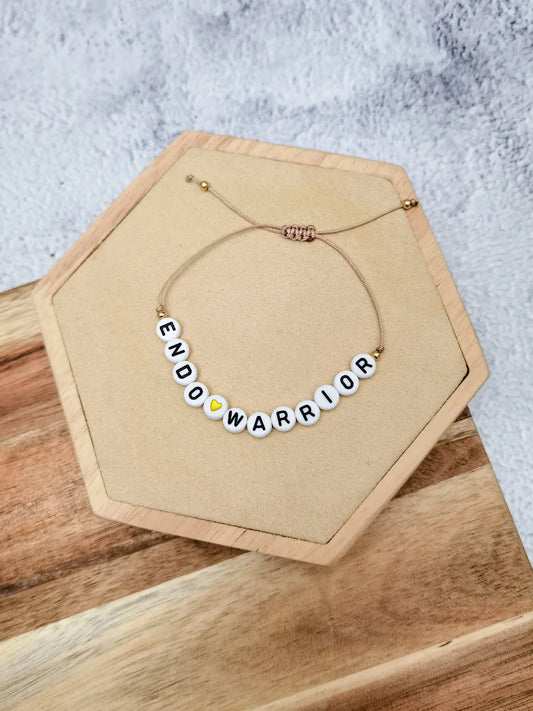 Beiges Endometriose Armband mit Buchstabenperlen &quot;Endo Warrior&quot; und einem goldenen Herz. Fotografiert auf einem Holz Sechseck, dass auf einem Holzbrett vor grauem Hintergrund liegt.