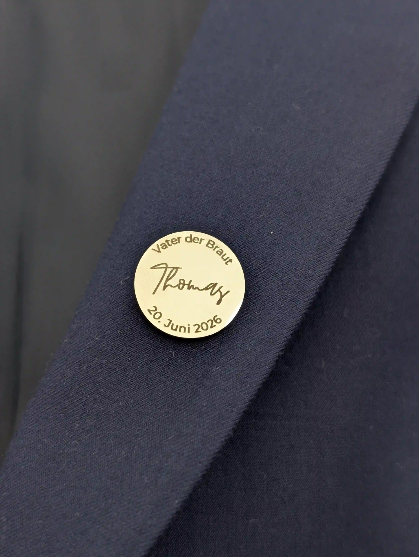 Personalisierter Anstecker in Gold am Revers eines dunkelblauen Anzugs, graviert mit „Vater der Braut", Name Thomas und Hochzeitsdatum 20. Juni 2026
