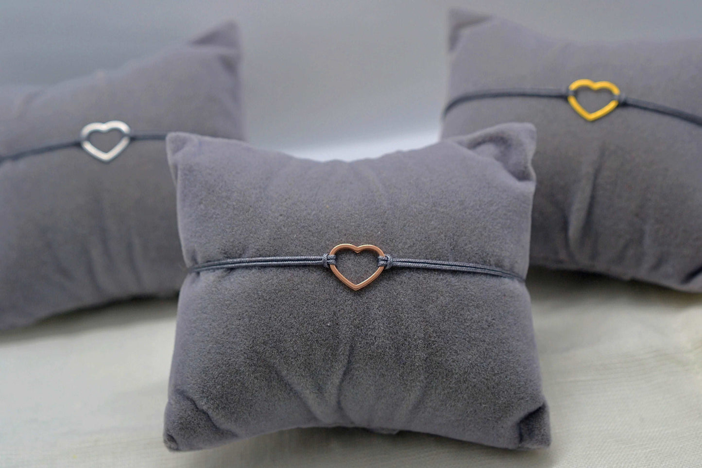 drei Herz Armbänder silber, gold und roségold mit grauem Band jeweils auf einem Schmuckkissen