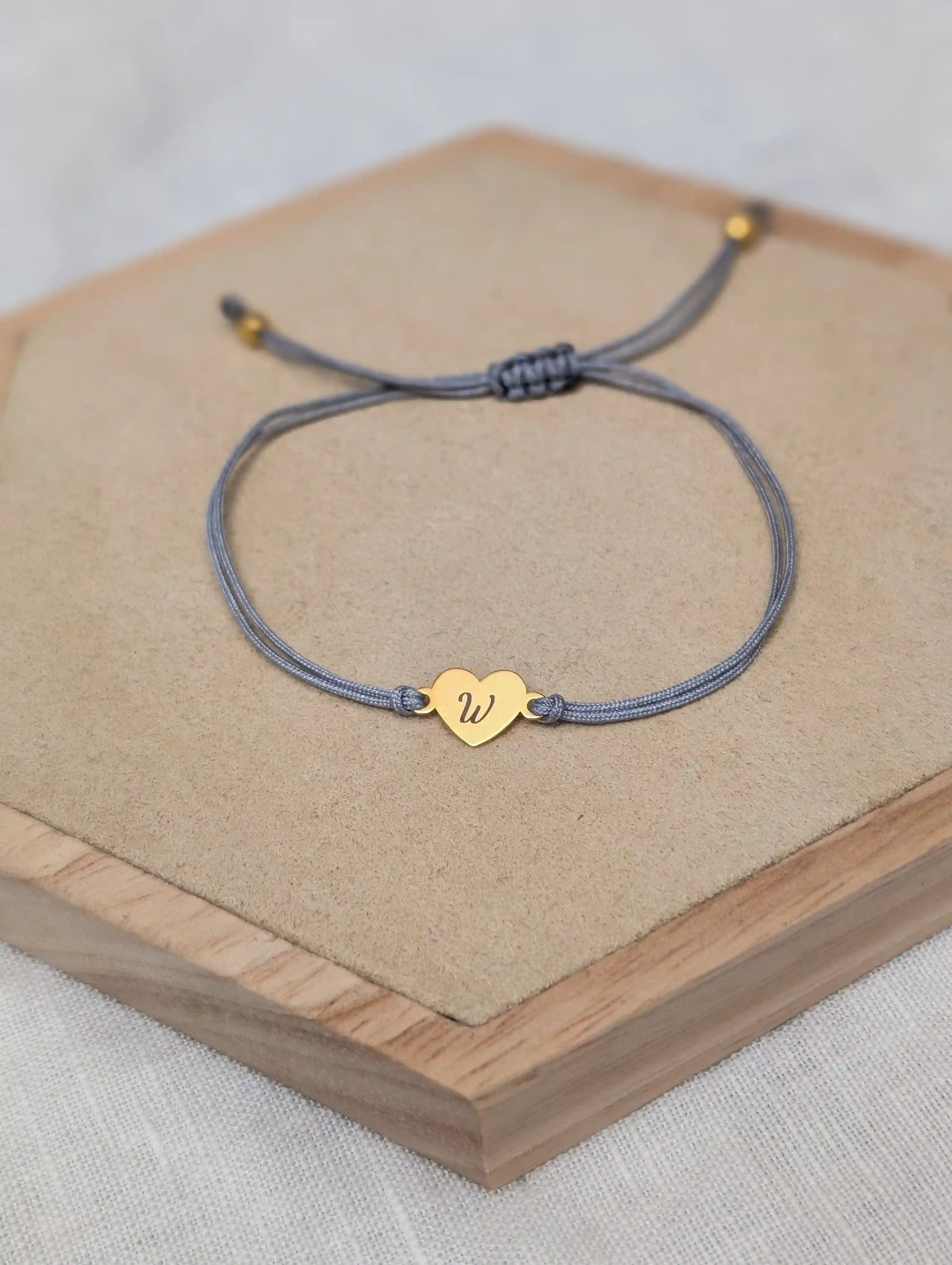 Ein graues Armband mit einem goldenen Herz Plättchen, auf das der Buchstabe "W" graviert ist, liegt auf einem Holz Sechseck