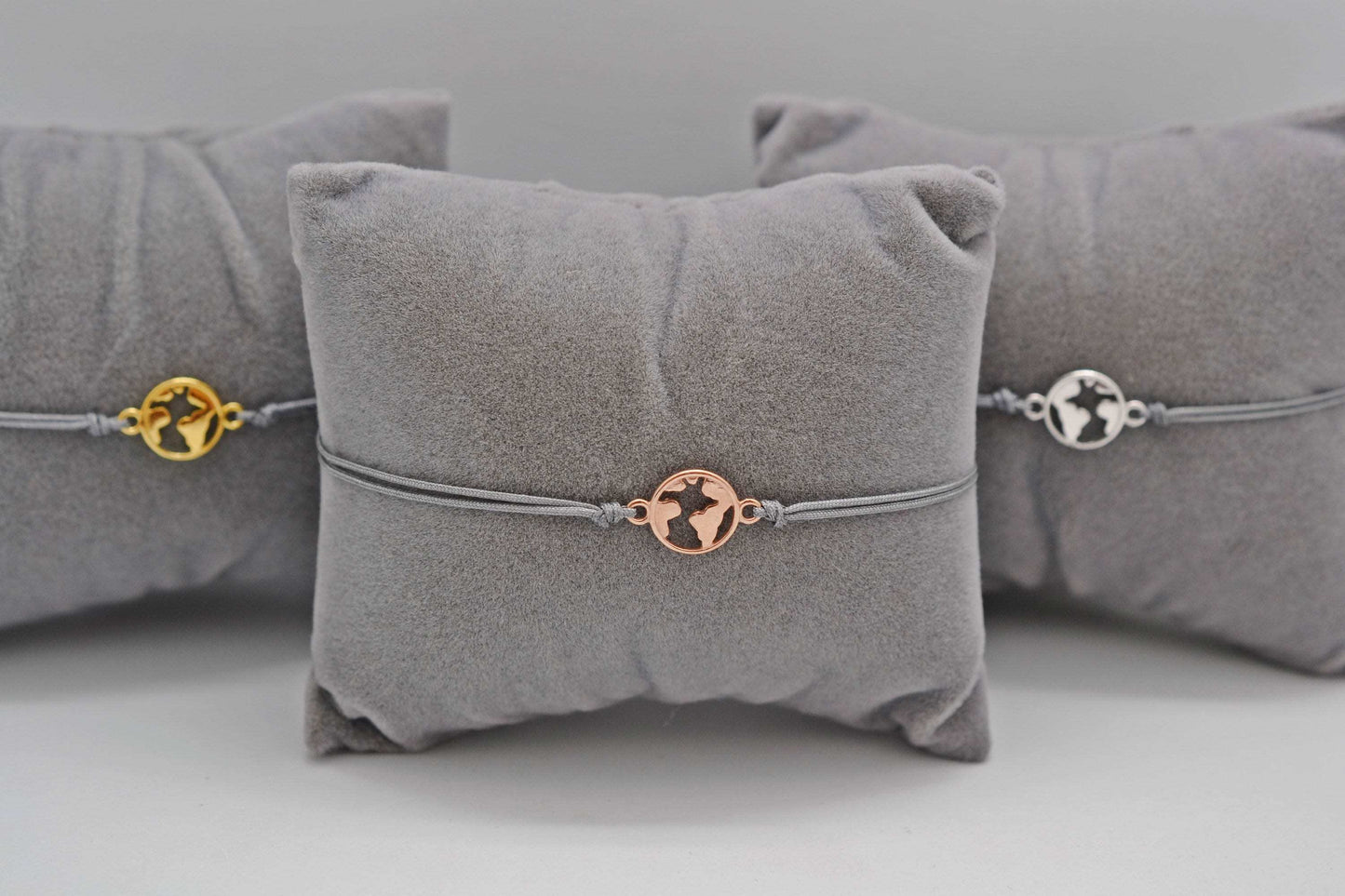 drei Armbänder mini Weltkarte in roségold, gold und silber mit grauem Band auf grauen Schmuckkissen