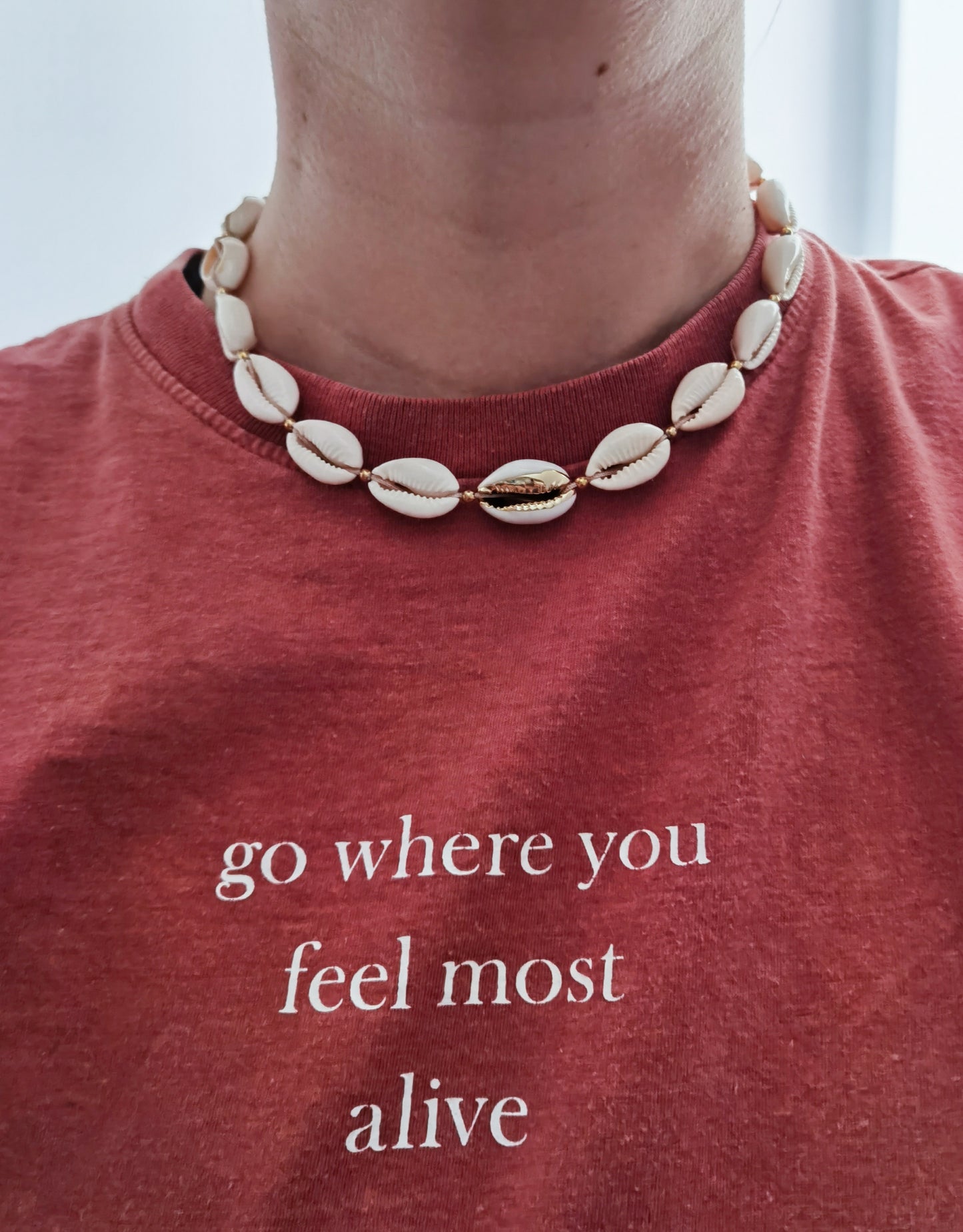 Nahaufnahme rotes T-Shirt mit Aufschrift "go where you feel most alive) und Muschelkette mit goldenen Akzenten