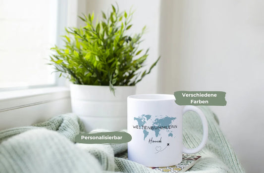 Weiße Tasse mit einer blauen Weltkarte und der Aufschrift Weltenbummlerin Hannah, im Hintergrund eine grüne Pflanze in weißem Topf