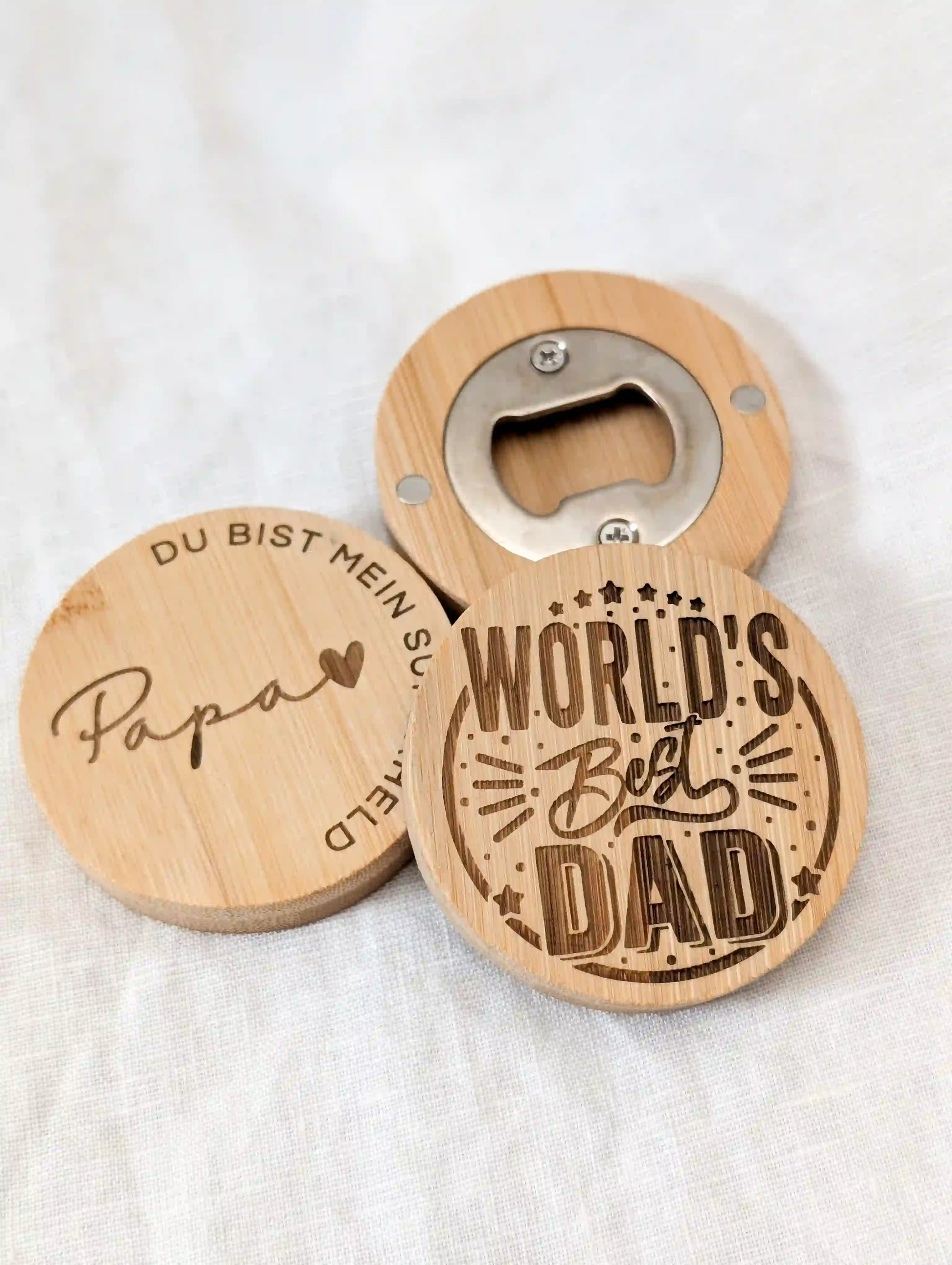 Drei runde Flaschenöffner aus Holz liegen auf einem weißen Tuch. Links Flaschenöffner für Papa mit Gravur "Papa du bist mein Superheld", schräg  davor Flaschenöffner zum Vatertag mit Gravur "World's best Dad" und hinten ein Öffner, der die magnetische Rückseite des Flaschenöffners zeigt