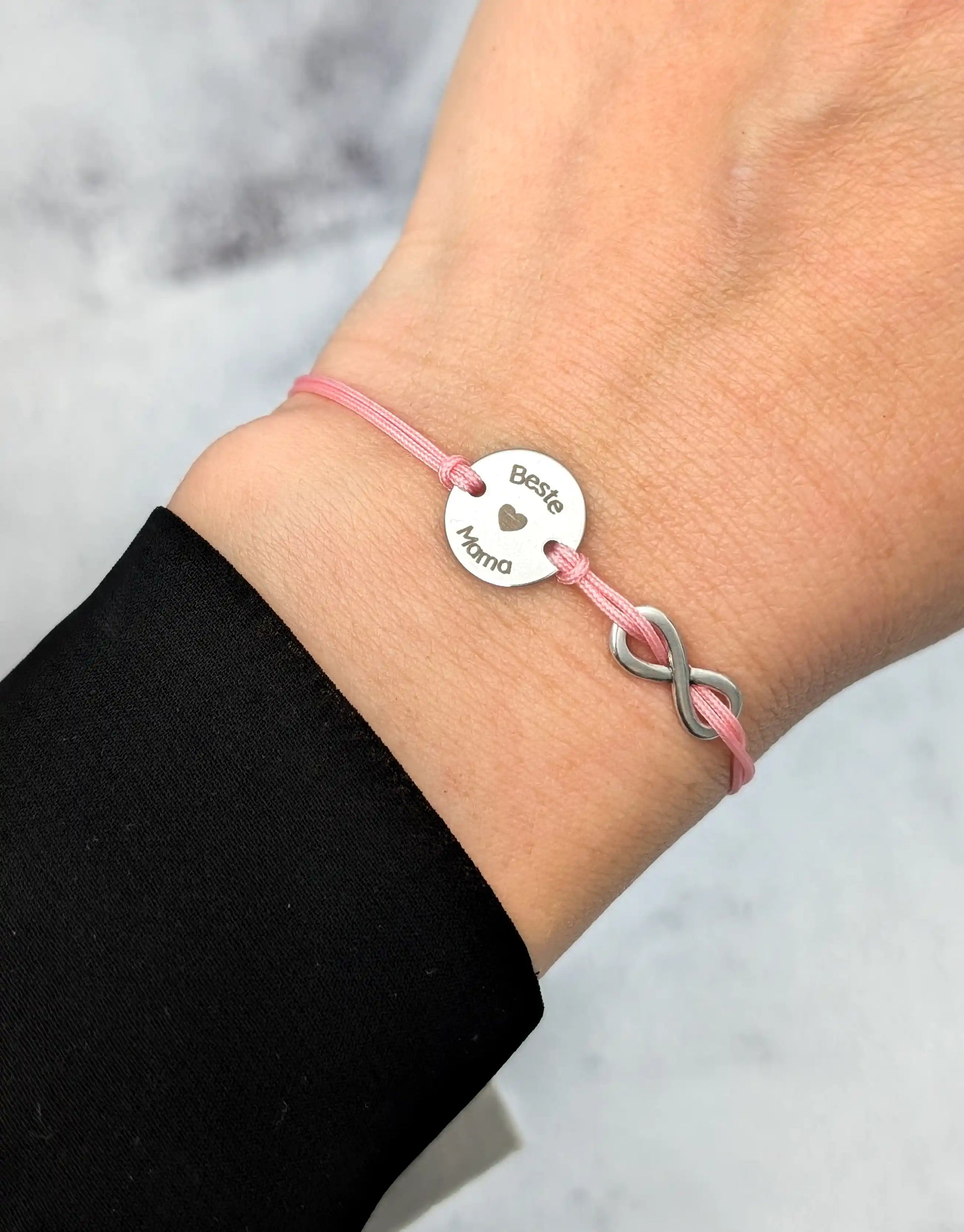 Tragefoto Rosafarbenes Gravur-Armband mit einem silbernen Plättchen mit Gravur "Beste Mama", daneben ein silbernes Unendlichkeitszeichen. 