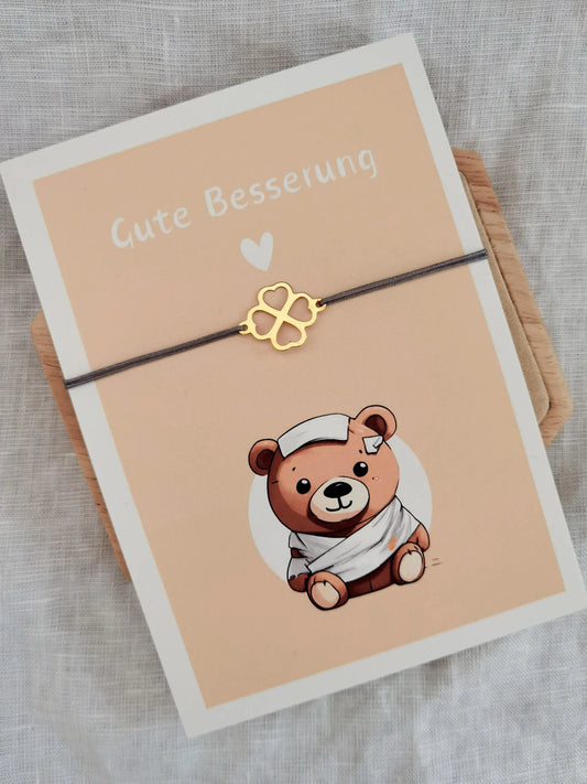 Postkarte Gute Besserung mit einem süßen Bären. Auf der Karte befestigt ein goldenes Kleeblatt Armband mit grauem Band