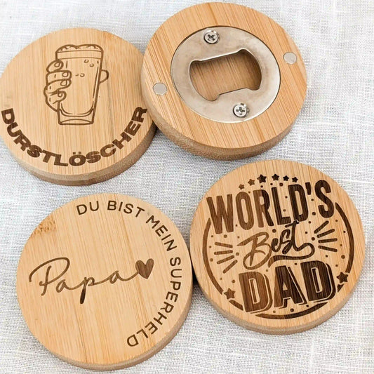 Vier runde Flaschenöffner aus Holz liegen in zwei Reihen auf einem weißen Tuch. Links oben Flaschenöffner mit Gravur "Durstlöscher", darunter "Papa du bist mein Superheld", daneben "World's best dad" und hinten ein umgedrehter Flaschenöffner, der die magnetische Rückseite zeigt