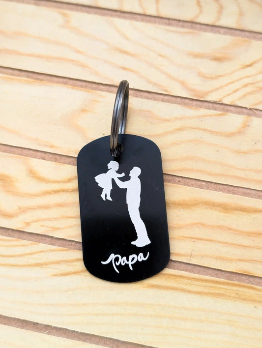 Ein schwarzer Schlüsselanhänger für Väter mit weißer Gravur eines Mannes der ein kleines Mädchen hochwirft. Darunter der Text "papa"