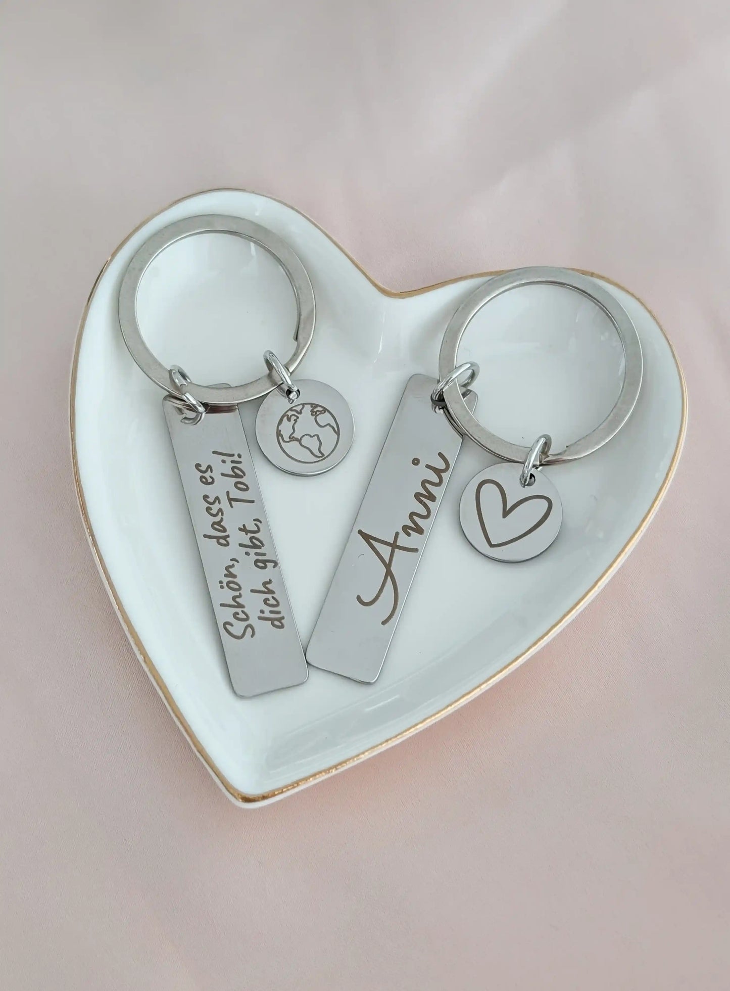 Zwei personalisierte Edelstahl-Schlüsselanhänger in einer herzförmigen Schale, graviert mit ‚Schön, dass es dich gibt Tobi‘ und einem Weltkarten-Symbol sowie ‚Anni‘ und einem Herzsymbol – individuelles Geschenk aus Edelstahl für besondere Menschen.
