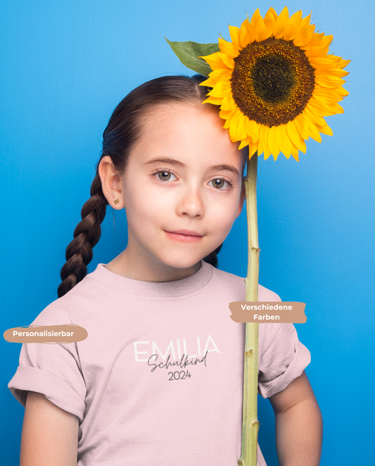 Mädchen mit geflochtenen Zopfen trägt ein personalisierbares T-Shirt &quot;Schulkind 2024&quot; in rosa. Mädchen hält eine große Sonnenblume in der Hand