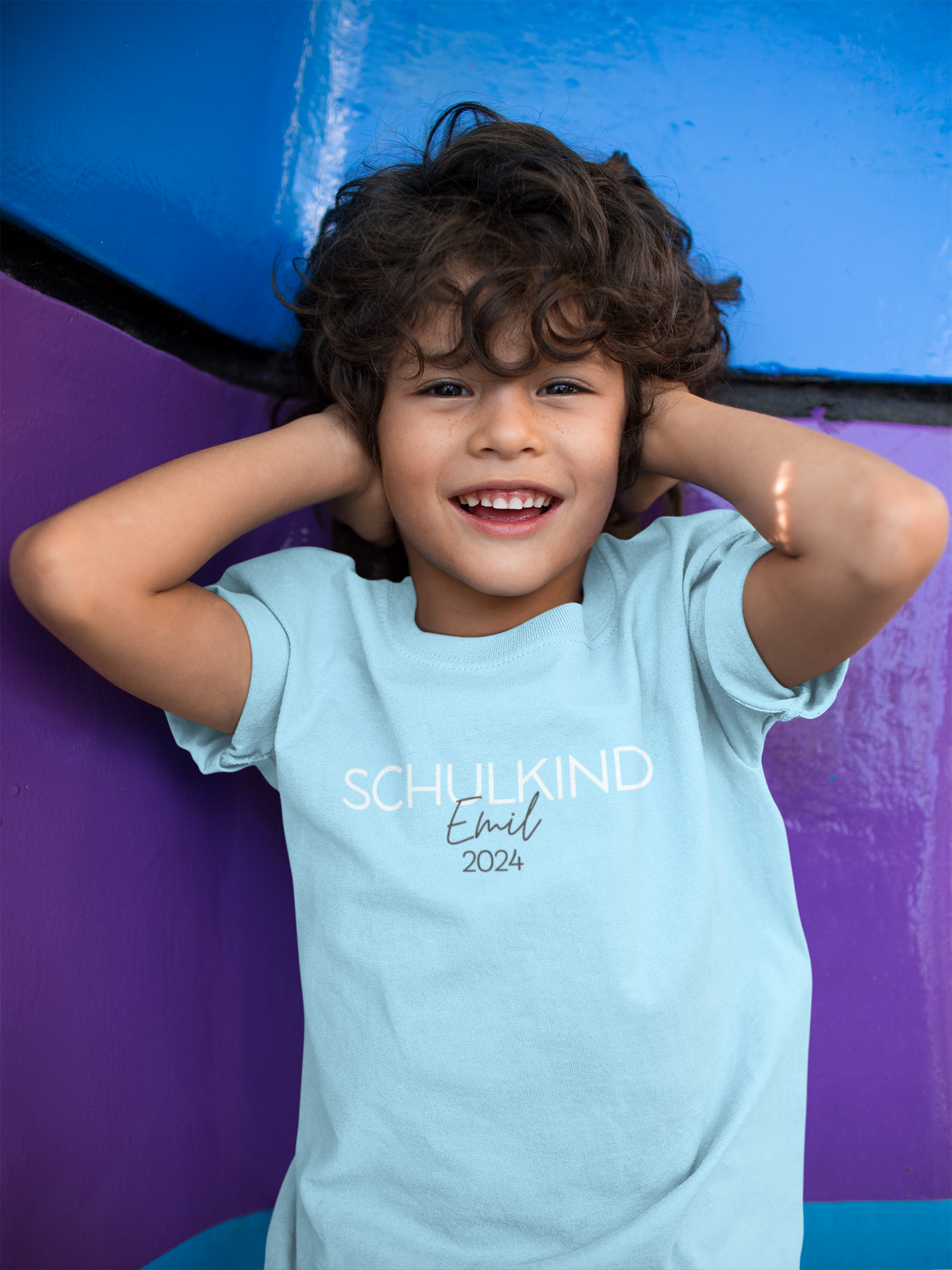 Ein Junge trägt ein personalisierbares T-Shirt "Schulkind 2024" in hellblau. Junge mit dunklen Haaren hält sich die Ohren zu