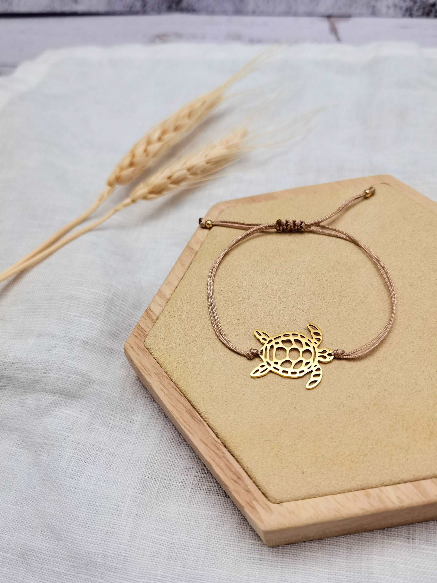 Armband mit einem goldenen Schildkröten Verbinder und beigem Band auf einem Holz Sechseck vor weißem Hintergrund