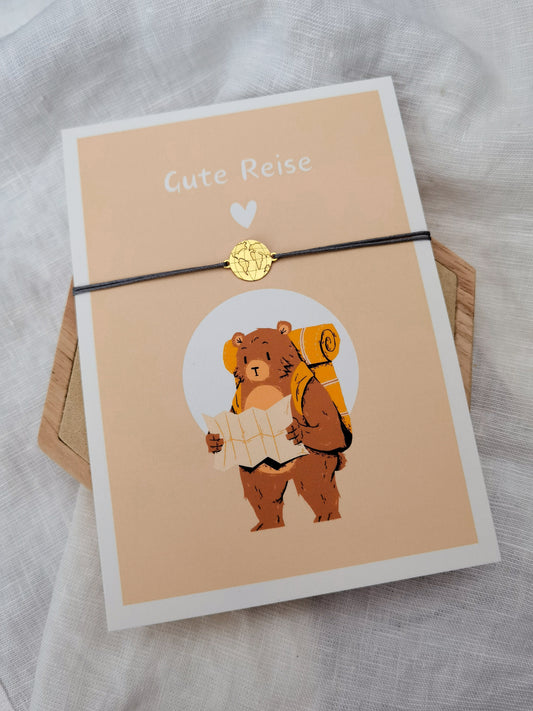 Postkarte mit der Aufschrift "Gute Reise" und einem Bären mit Rucksack und Landkarte in der Hand. Weltkarten Armband in gold und grau an der Karte befestigt
