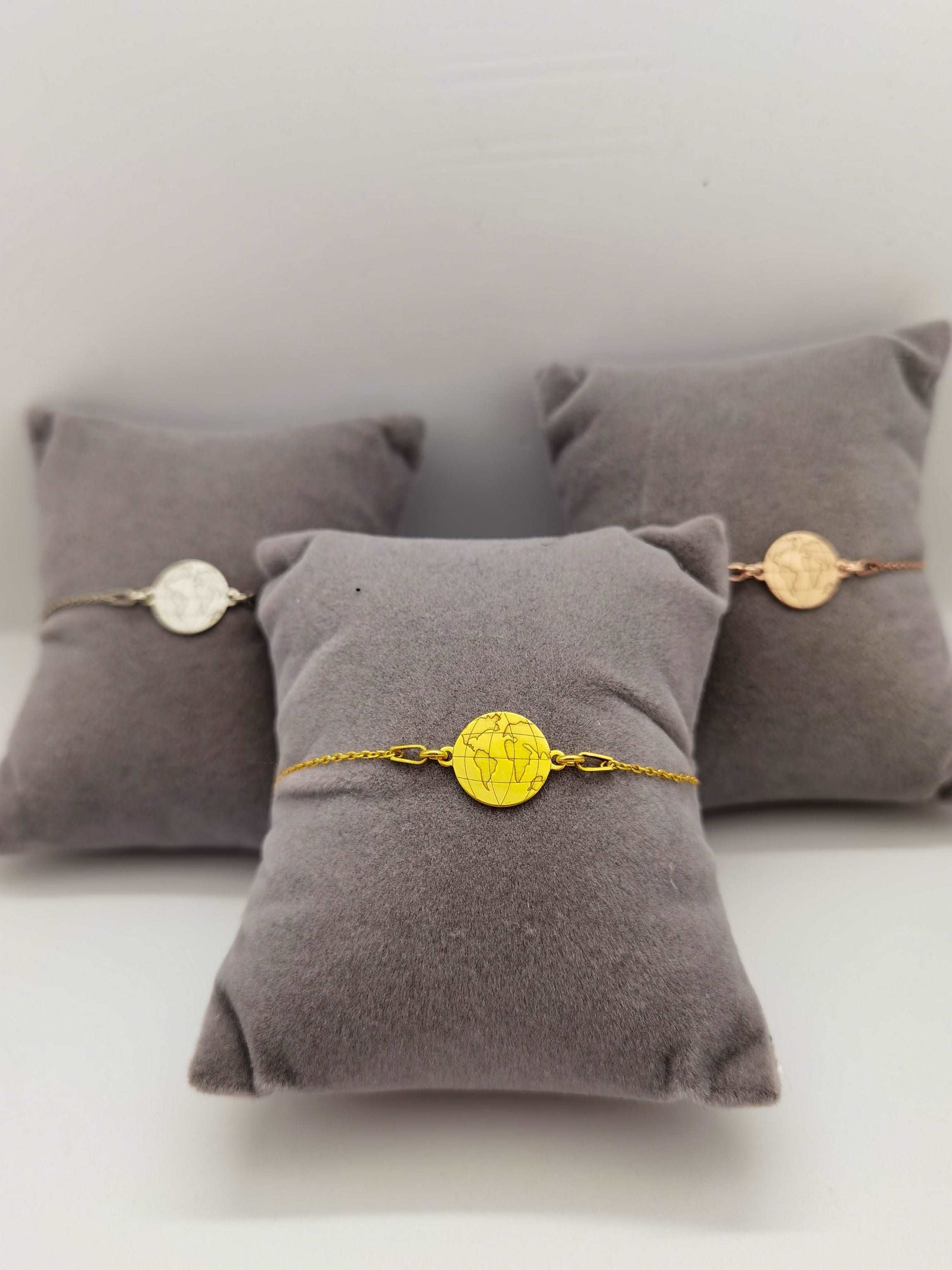 Drei graue Schmuckkissen mit Weltkarten Armbändern in silber, gold und roségold. Goldenes Armband im Fokus